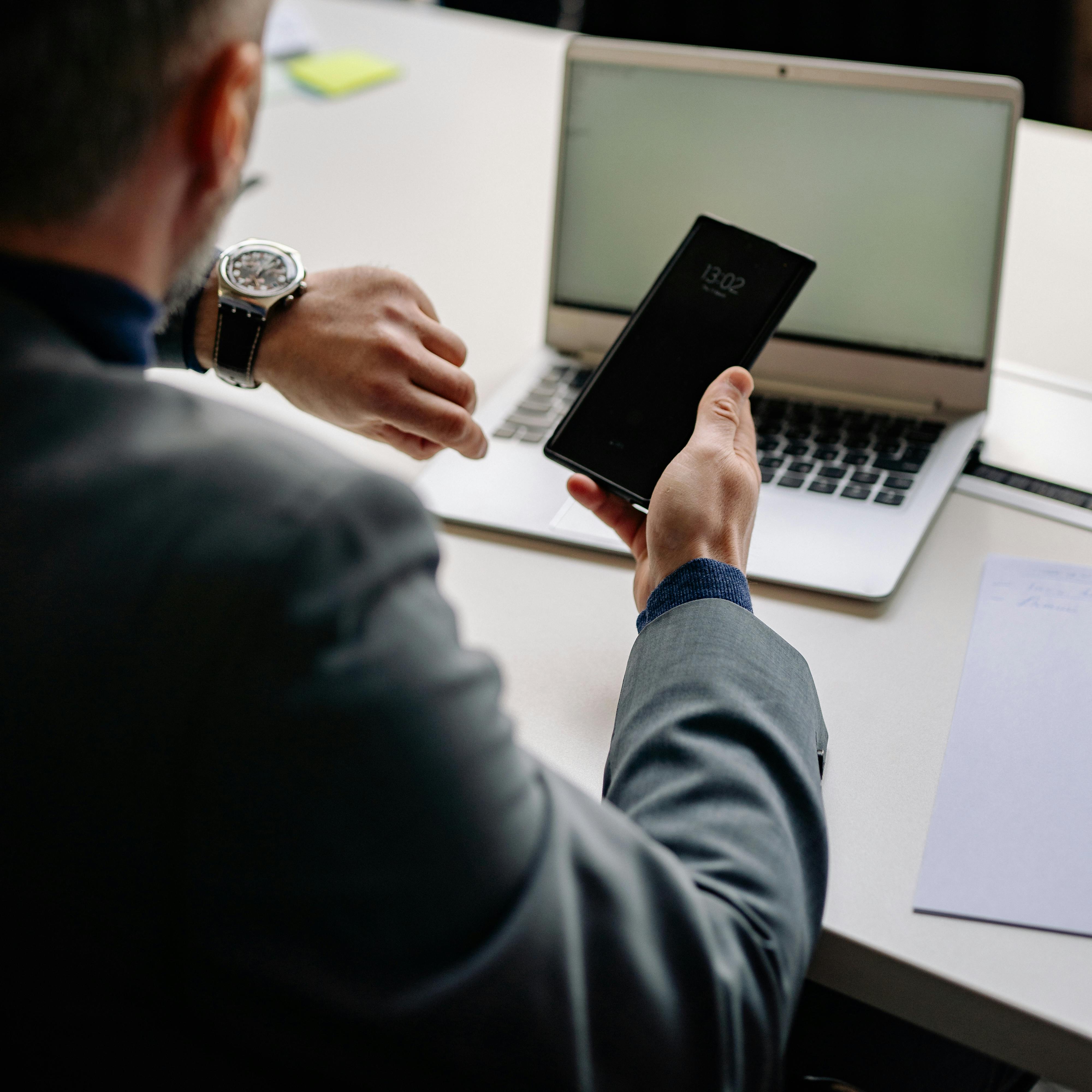 Man in office using smartphone and laptop, showing time management.